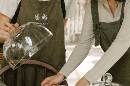 Food Careers - Positive female colleagues wearing aprons standing at table with glass dome lids and various desserts while working in modern cafeteria
