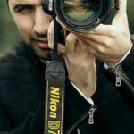Career Exploration - Concentrated male photographer in casual clothes looking through objective lens of camera while taking photo on nature with blurred background