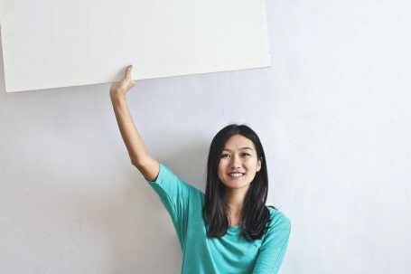 Real Estate Career - Cheerful Asian woman sitting cross legged on floor against white wall in empty apartment and showing white blank banner