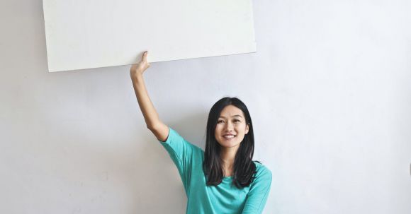 Real Estate Career - Cheerful Asian woman sitting cross legged on floor against white wall in empty apartment and showing white blank banner